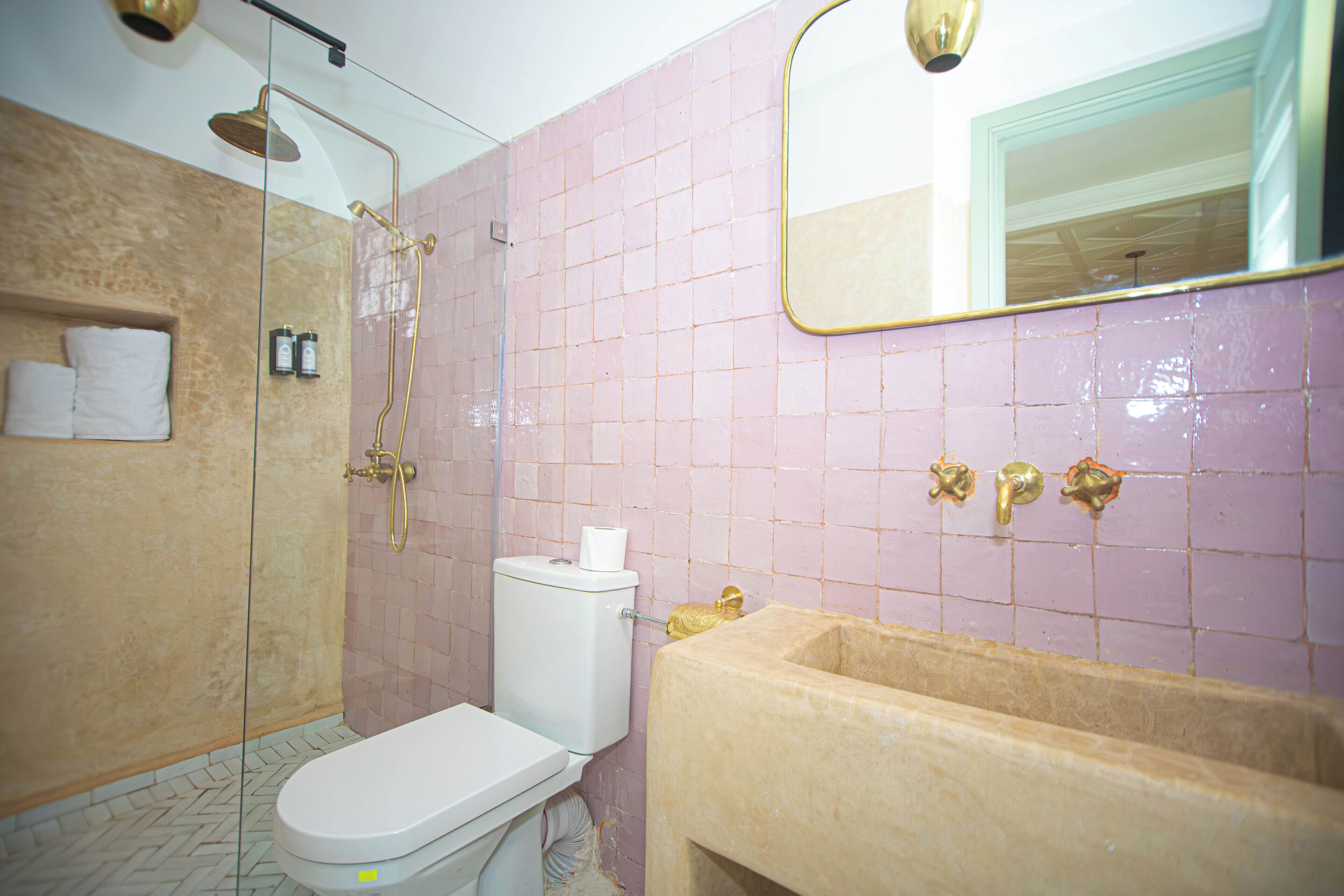 Baño con inodoro, ducha y lavamanos, con azulejos rosa y accesorios dorados.