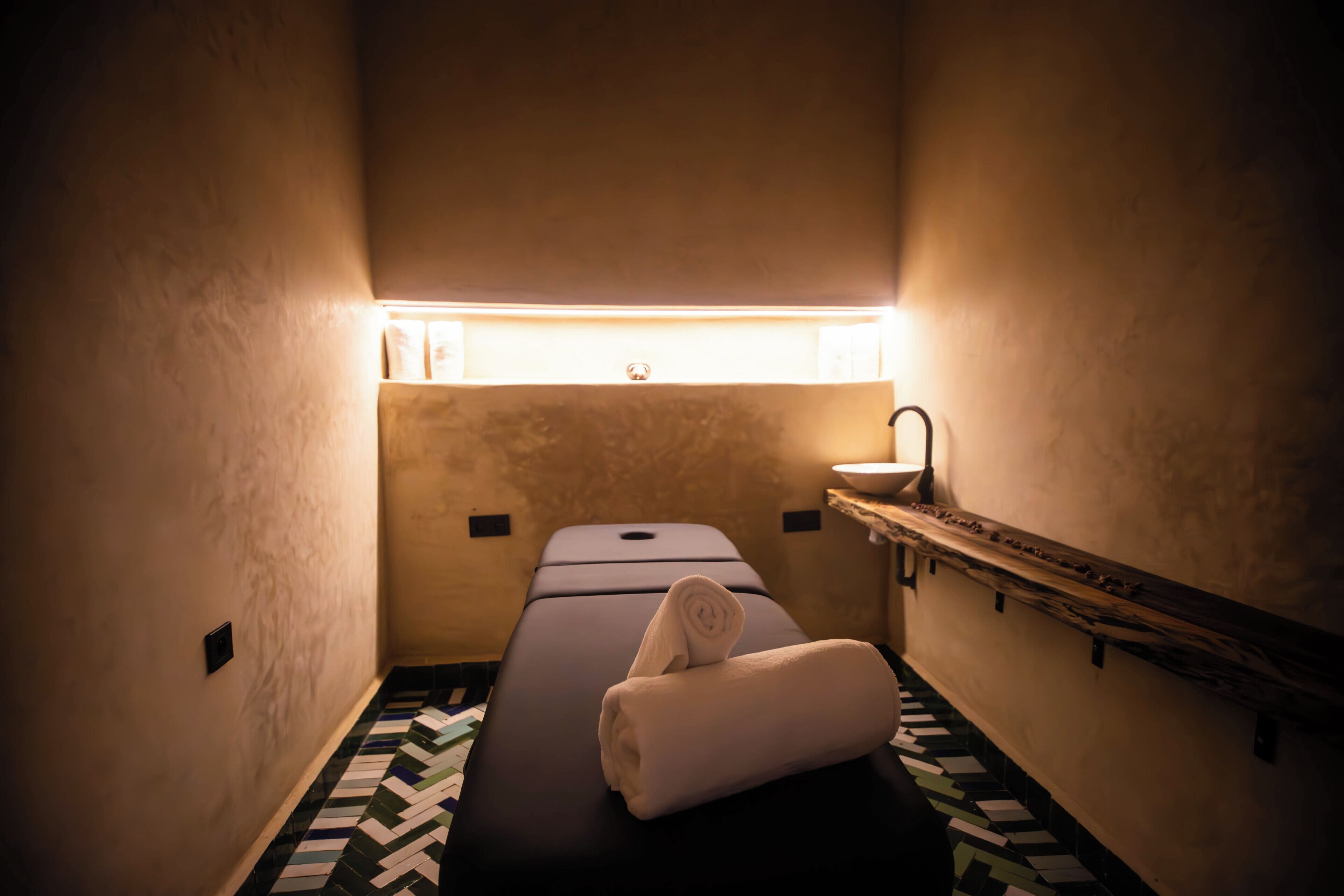 A small, dimly lit room with a single bed, a small sink, and a patterned floor.