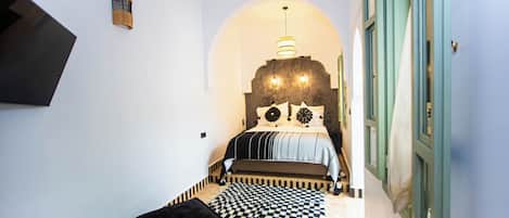 Un pasillo estrecho que conduce a un dormitorio con una cama, una pequeña mesa redonda, un tapete a cuadros blanco y negro y un televisor montado en la pared.