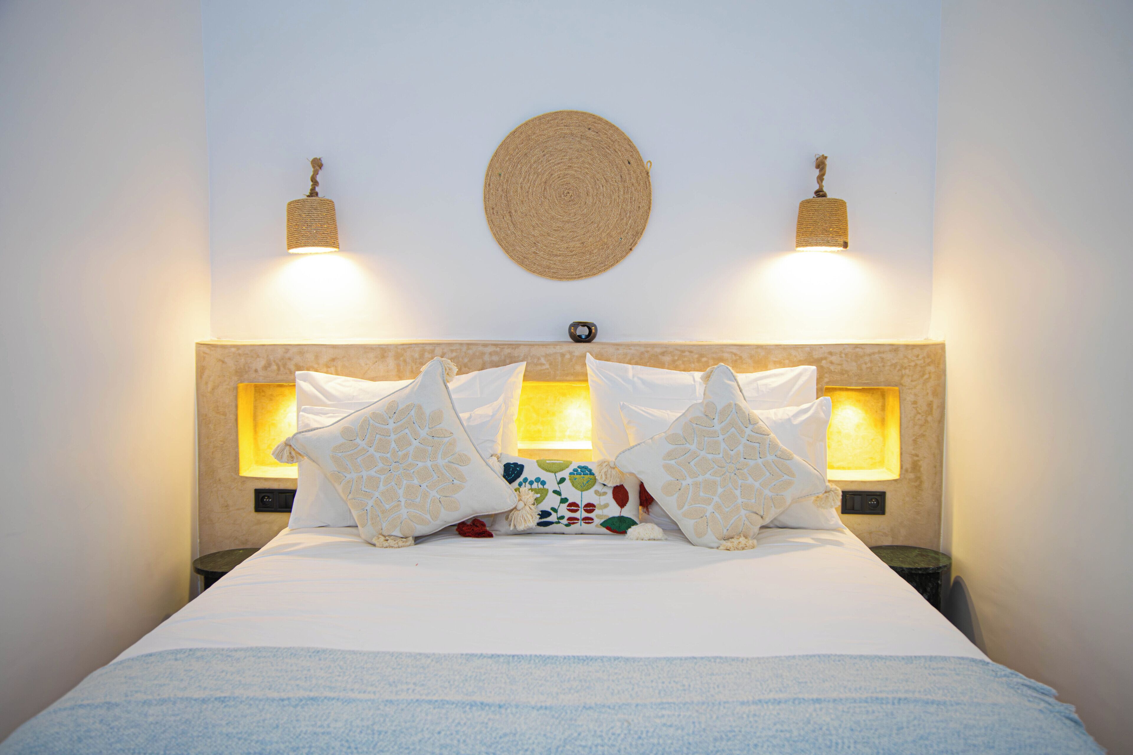 A neatly made bed with white linens, two wall-mounted lamps, and a woven wall hanging.