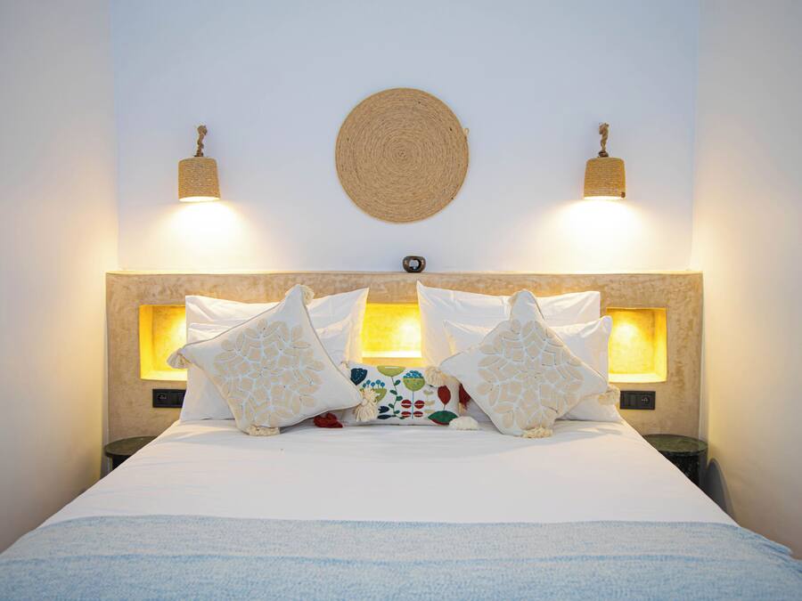 A neatly made bed with white linens, two wall-mounted lamps, and a woven wall hanging.