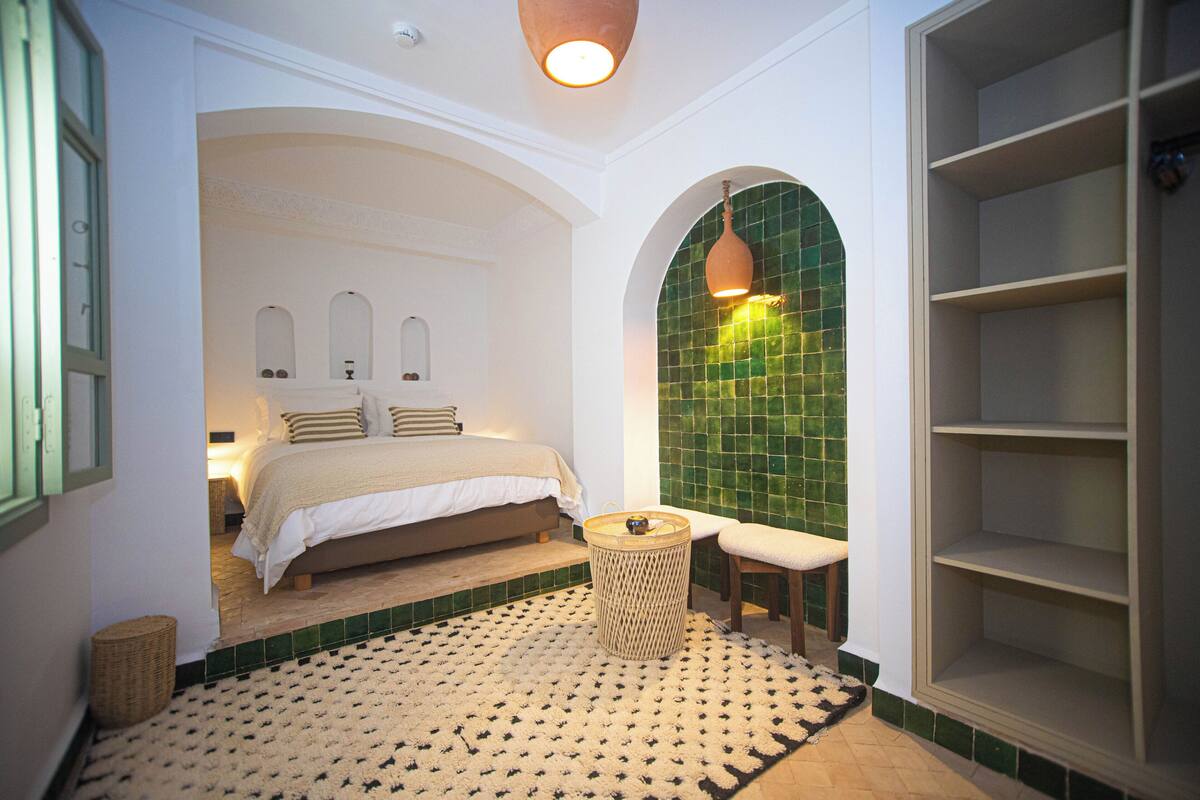 Ein Schlafzimmer mit einem Bett, einem kleinen runden Tisch, einer Bank und einer grünen Wandfliese.