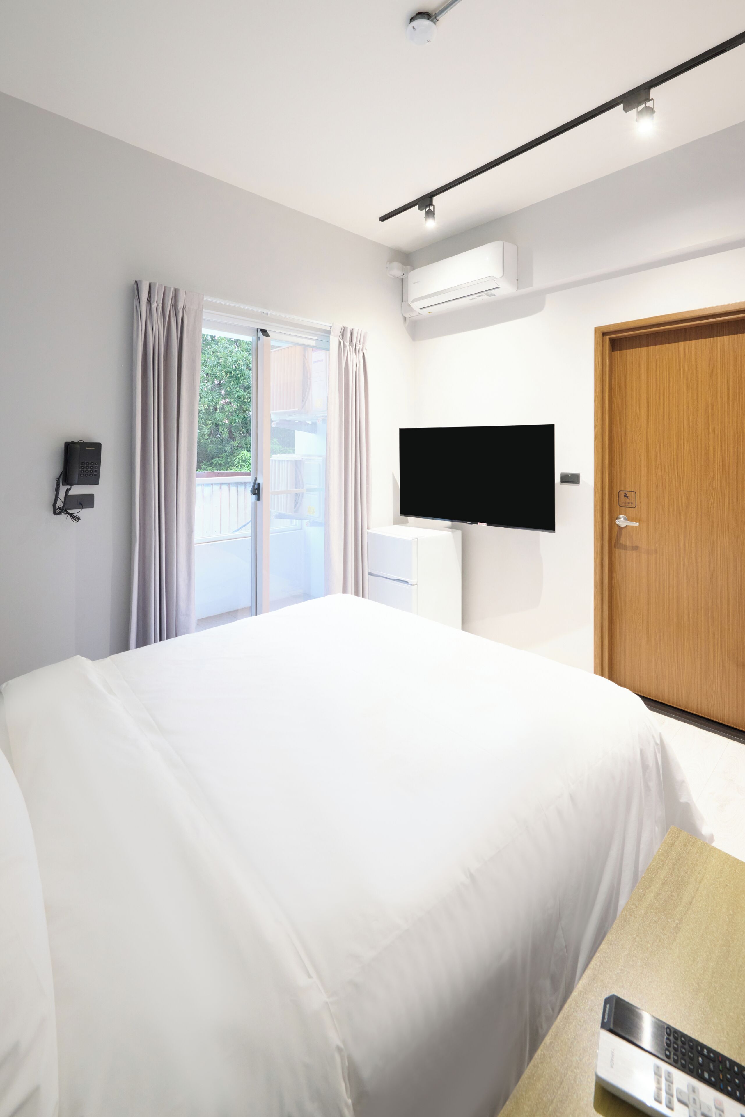 A hotel room with a bed, a TV mounted on the wall, an air conditioning unit, a telephone, and a wooden door.