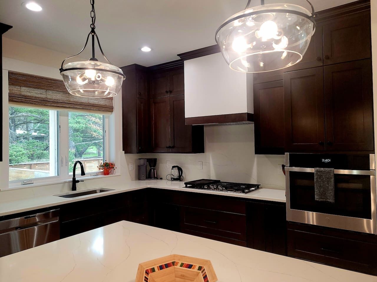 A modern kitchen with dark wood cabinets, a white countertop, a stainless steel oven, and a pendant light fixture.