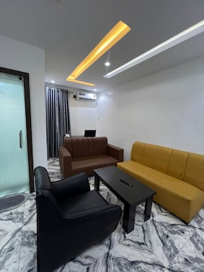 Moderni hotellihuone, jossa on marmorilattia, ruskea sohva, keltainen sohva, musta nojatuoli ja musta sohvapöytä.