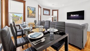 A modern dining area with a long table, chairs, and a television displaying 'Roku.' - The Milton Manor 5BR 2BA - Sleeps 10 - Unit 2 (Milton)