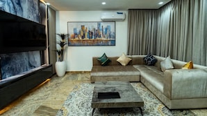 Una sala moderna con un sofá seccional, un televisor de pantalla plana, un cuadro de un paisaje urbano y una planta grande en maceta.
