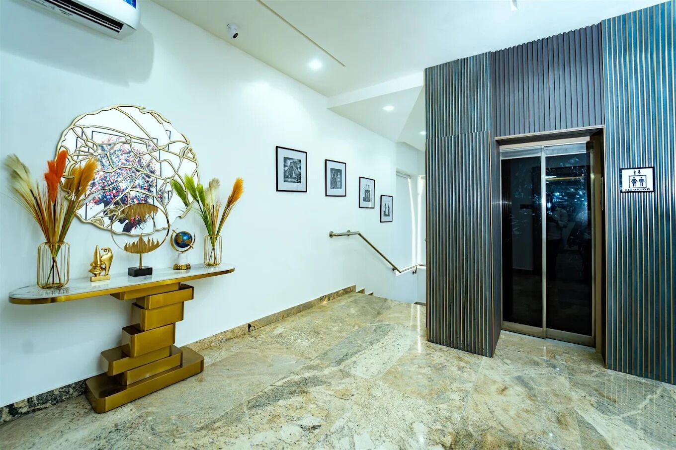 A hotel lobby with a marble floor, a reception desk with a gold finish, framed pictures on the wall, and a modern elevator.
