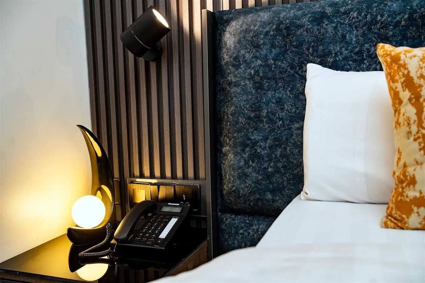 Una habitación de hotel con una cama, una mesita de noche con lámpara, un teléfono y una lámpara de pared.