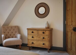 Un dormitorio con un sillón capitonado, un aparador de madera con cajones, un espejo redondo y una puerta de madera.