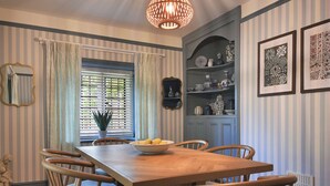 A dining area with a wooden table and chairs, a bowl of lemons, striped wallpaper, a mirror, and a hanging light fixture. - Home Farm Cottage (Peterborough)