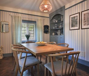 A dining area with a wooden table and chairs, a bowl of lemons, striped wallpaper, a mirror, and a hanging light fixture. - Home Farm Cottage (Peterborough)