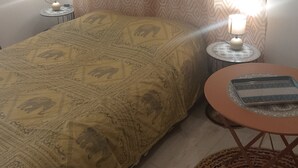 Ein Schlafzimmer mit einem Bett, zwei Nachttischlampen, einer gemusterten Tagesdecke, einem kleinen runden Tisch mit Tablett und einem Vorhang.