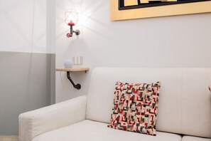 Un canapé blanc agrémenté d’un coussin aux motifs géométriques colorés, un luminaire fixé au mur et une petite étagère ornée d’objets décoratifs.