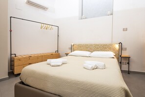 Un lit bien fait, recouvert d’une courtepointe beige, avec deux oreillers et deux serviettes enroulées. On y trouve également un banc en bois avec des tiroirs, un porte-manteaux fixé au mur et une petite table avec une lampe.