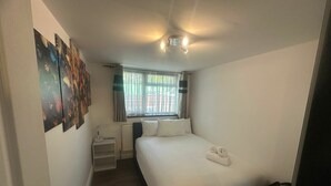 A compact hotel room with a single bed, a small bedside table, a radiator, and a window with curtains. - Modern 2 BDR APT Havelock Road UB2 (Greater London)