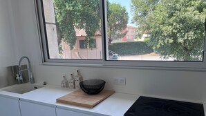 A modern kitchen with a white countertop, a black induction cooktop, a wooden cutting board, and a black woven bowl. - 🏡 Bright apartment in Fréjus - beach, town center, night market (Fréjus)