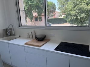 A modern kitchen with a white countertop, a black induction cooktop, a wooden cutting board, and a black woven bowl. - 🏡 Bright apartment in Fréjus - beach, town center, night market (Fréjus)