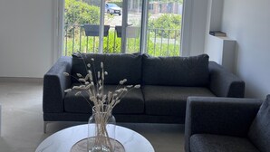A modern living room with a grey sofa, a round marble table, and a view of a residential area through large windows. - 🏡 Bright apartment in Fréjus - beach, town center, night market (Fréjus)