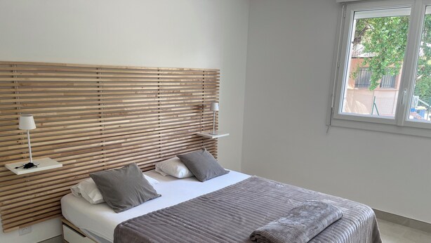 A modern bedroom with a wooden headboard, a bed with white and gray bedding, a bedside table with a lamp, and a window with a view of greenery. - 🏡 Bright apartment in Fréjus - beach, town center, night market (Fréjus)