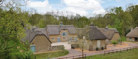 A row of traditional thatched-roof cottages with stone walls, surrounded by a fenced pathway and lush greenery.