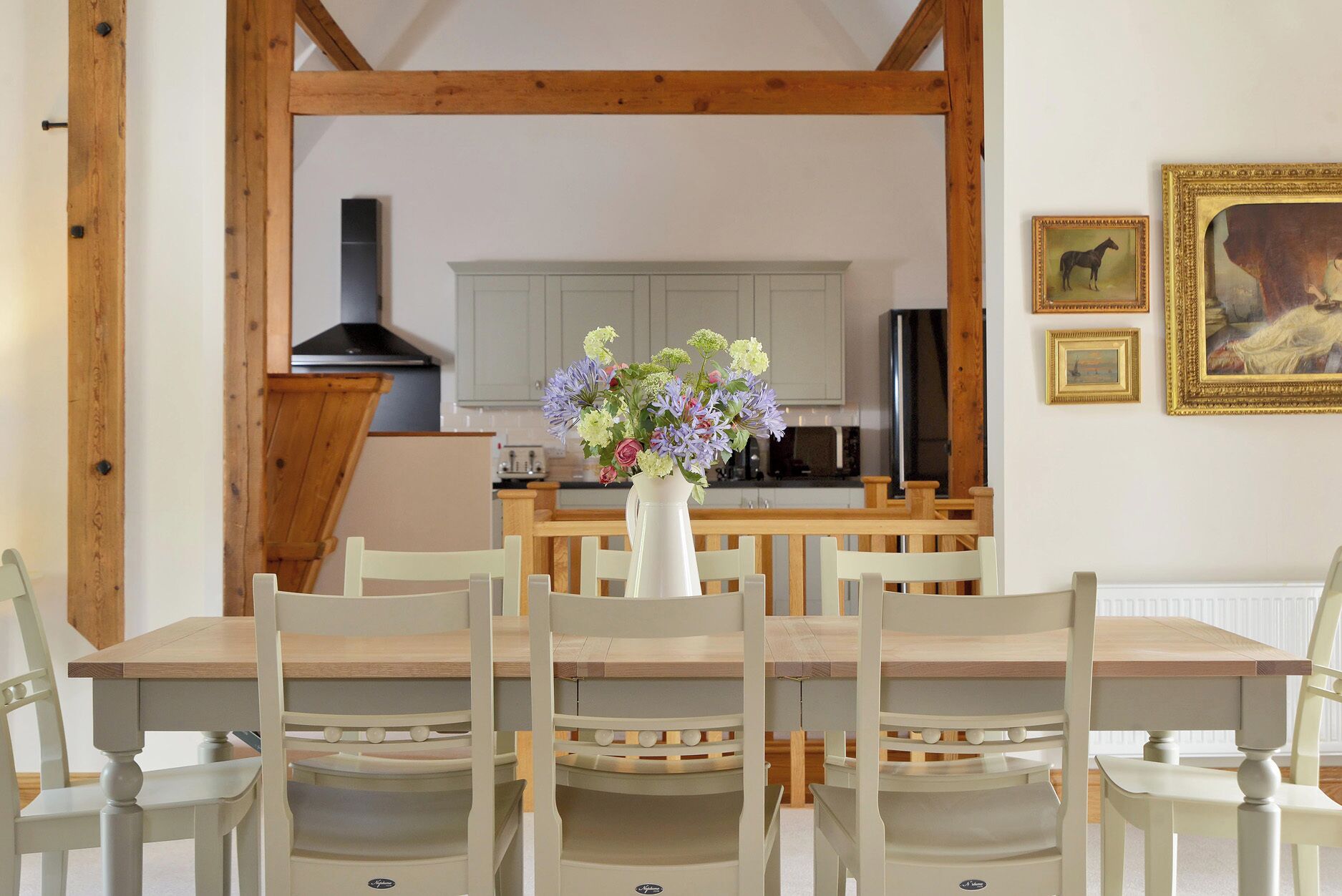 A dining area with a table and chairs, a vase with flowers, a kitchen in the background, and framed pictures on the wall.