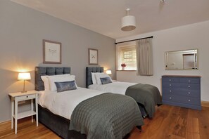 A bedroom with two beds, a nightstand, a dresser, a window with curtains, and a ceiling light. - The Lady Rothschild Dairy (Peterborough)