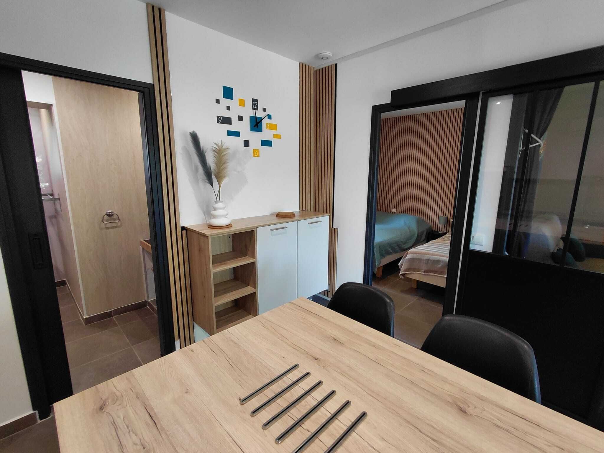 Een moderne kamer met een houten eettafel, een kleine kitchenette, een bed en een glazen schuifdeur naar een andere kamer.