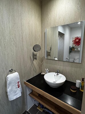 Un baño con un lavamanos blanco, un espejo, un perchero para toallas y un estante pequeño con artículos de aseo.