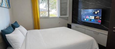 A hotel room with a bed, a TV, a window with curtains, a remote control, and a painting on the wall.