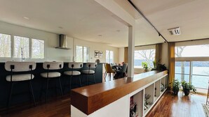 A modern kitchen with a bar area, wooden flooring, and large windows overlooking a body of water. - The Grand Lakeview – Midcentury Modern Villa (Ithaca)