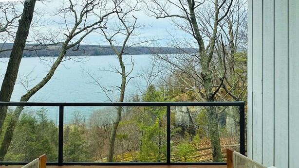 A balcony with wicker furniture, a bottle, and a glass, overlooking a lake and trees. - The Grand Lakeview – Midcentury Modern Villa (Ithaca)