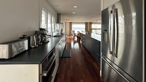 A modern kitchen with stainless steel appliances, a wooden floor, and large windows offering a view. - The Grand Lakeview – Midcentury Modern Villa (Ithaca)