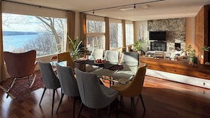 A modern living room with a dining area, large windows, a stone fireplace, and a television. - The Grand Lakeview – Midcentury Modern Villa (Ithaca)
