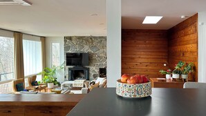A modern living room with a stone fireplace, wooden paneling, and a dining area with a table and chairs. - The Grand Lakeview – Midcentury Modern Villa (Ithaca)