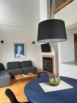 A modern living room with a grey sofa, a coffee table, a dining table with a blue tablecloth, a black lamp, a fireplace, and a framed artwork on the wall.