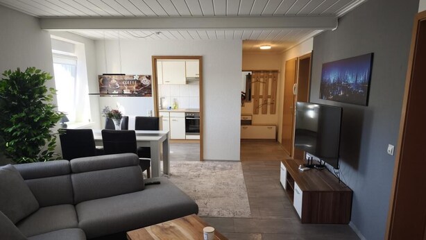 En moderne stue med en hjørnesofa, et træsofabord, et fjernsyn og et køkken i baggrunden.
