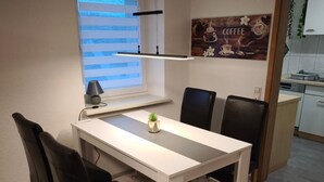 Et spiseområde med et hvidt bord, sorte stole, en lille plante og et køkken i baggrunden.