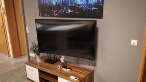 En moderne stue med et fladskærms-tv monteret på væggen, et træunderholdningsmøbel med en vase med blomster, og et bybillede som billede over tv'et.