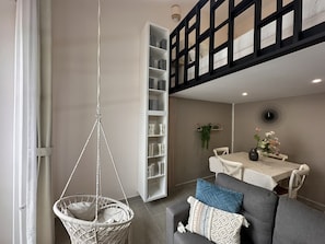 Een kamer met een hangstoel, een eettafel met stoelen, een boekenkast en een raam met een zwarte lijst.