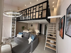 Een moderne woonkamer met een grijze bank, een glazen salontafel, een eethoek met een witte tafel en stoelen, een trap en een boekenkast.