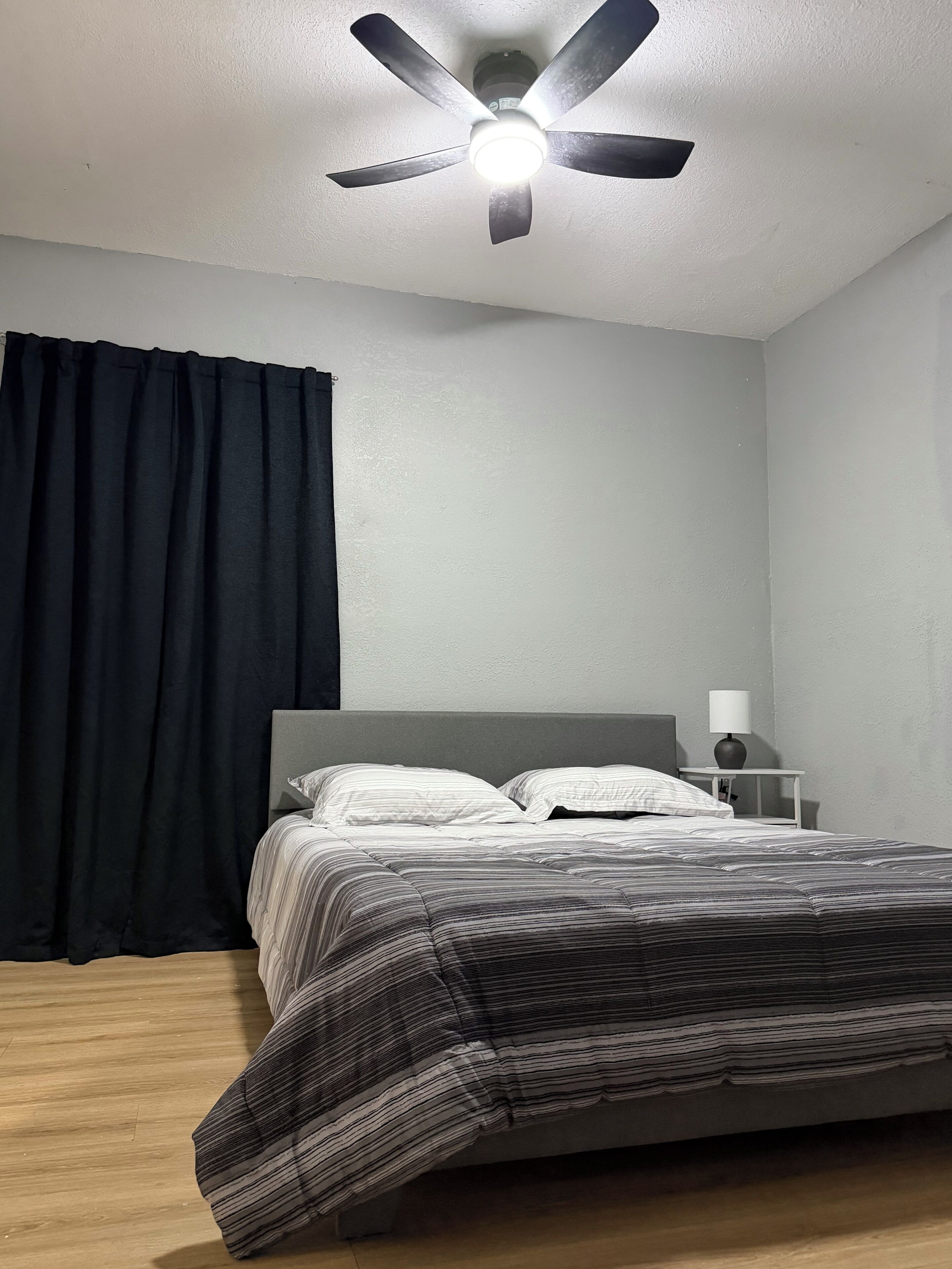 A bedroom with a bed, a nightstand, a lamp, a ceiling fan, and a window with curtains.