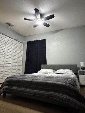 Ein Schlafzimmer mit Bett, Deckenventilator, Nachttisch mit Lampe, Fenster mit Vorhängen und Kleiderschrank.