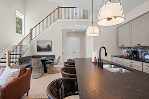 A modern kitchen with a central island, a dining area with wicker chairs, a staircase, and a view of the outdoors. - 4BR Near the Ark Encounter (Dry Ridge)