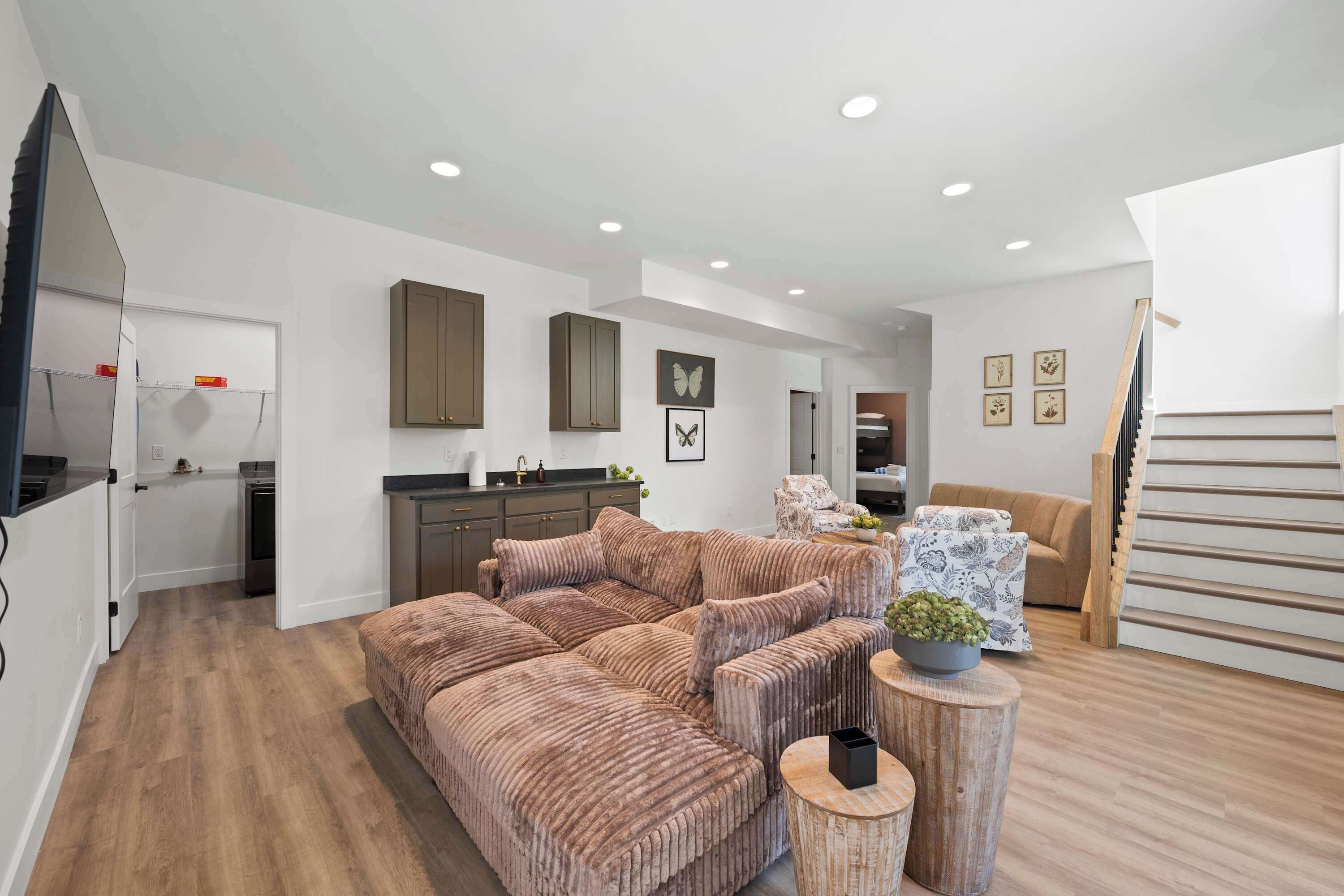 A modern living room with a sectional sofa, a dining area with a kitchen in the background, and a staircase leading to an upper level.