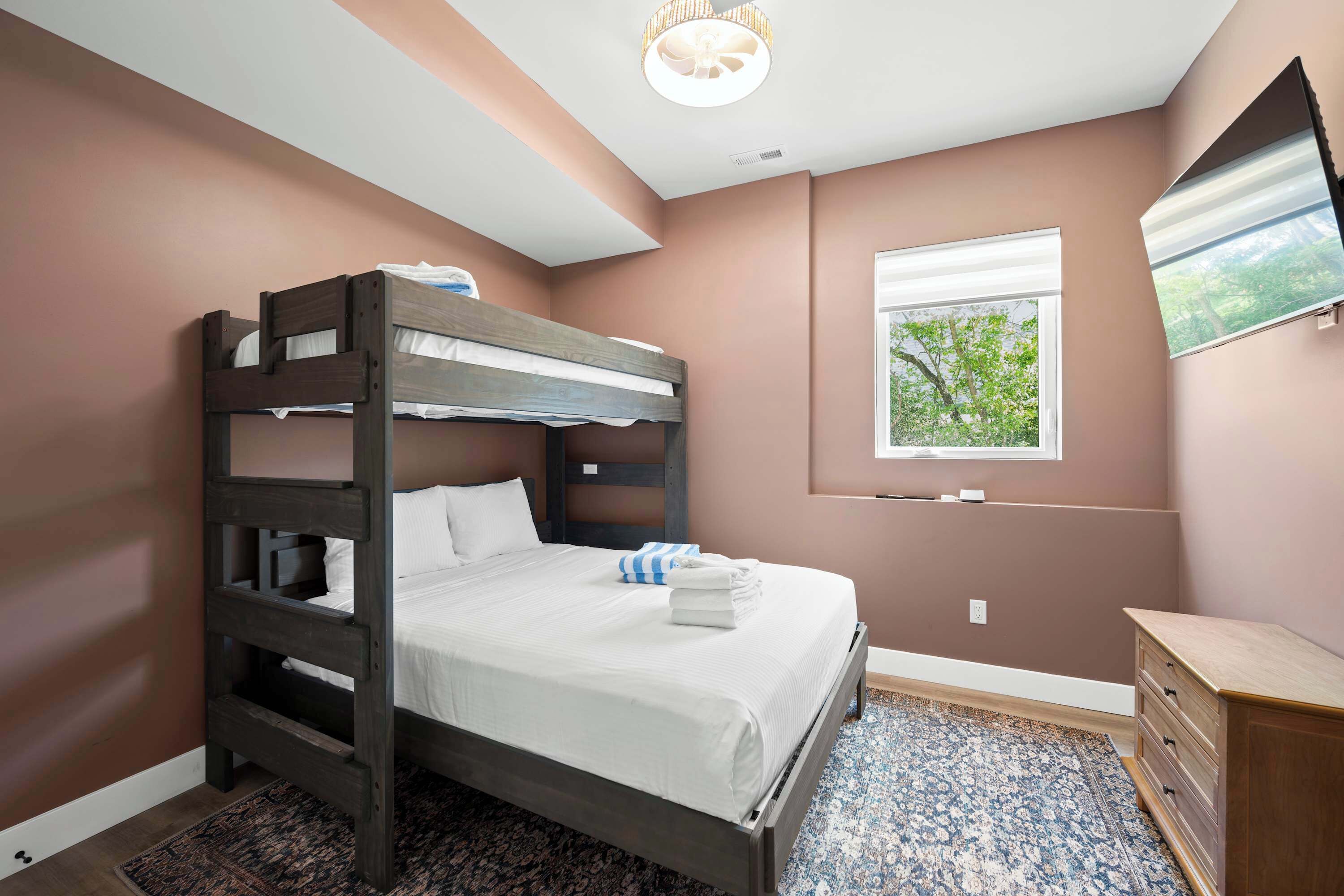 A bunk bed with a window, a dresser, and a patterned rug in a room with a brown wall and ceiling light.