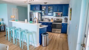 Una cocina con armarios azules, un refrigerador de acero inoxidable, un microondas, un horno y un lavamanos. Hay tres taburetes azul claro y un bote de basura.