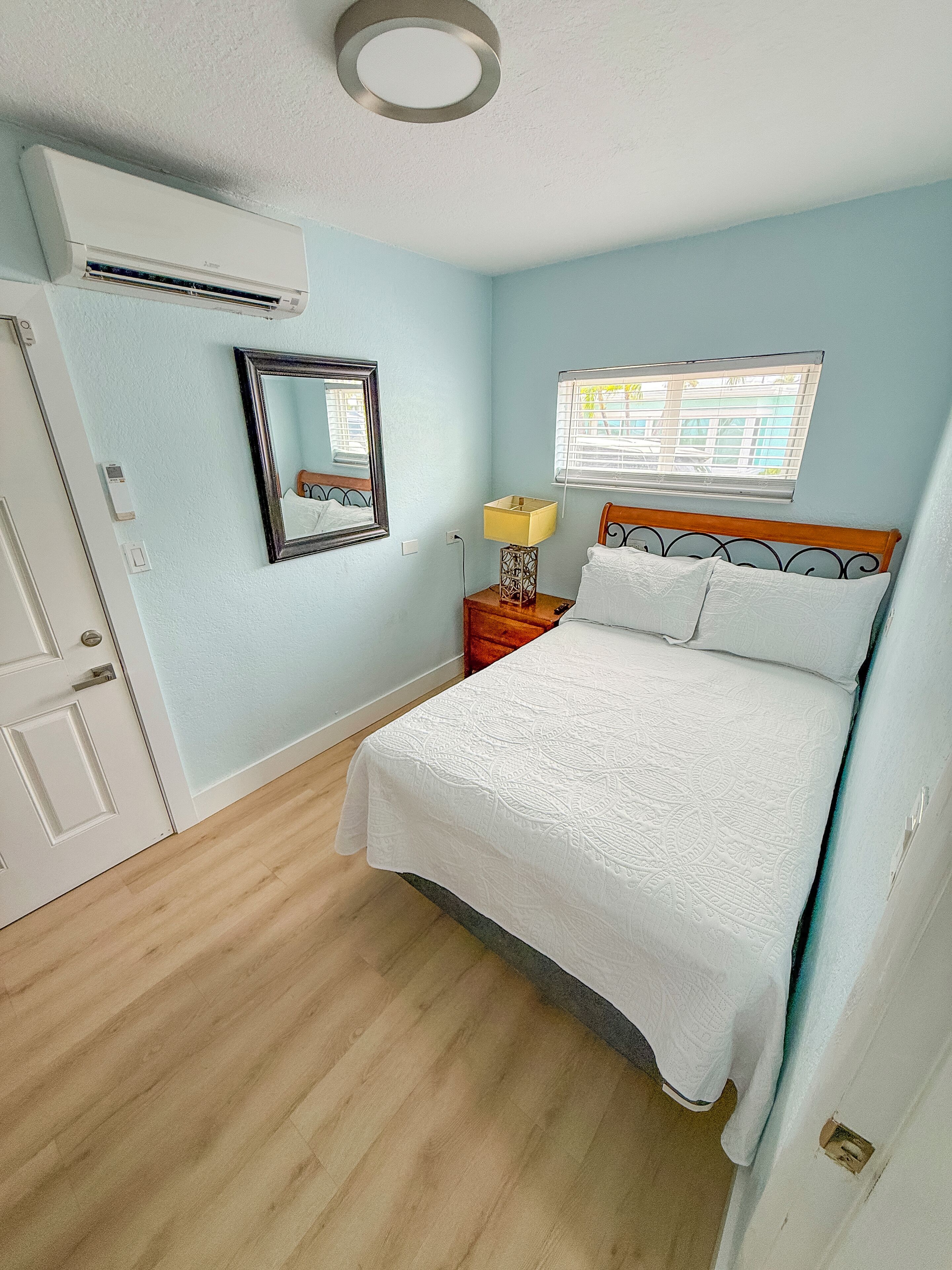 Une chambre à coucher comprenant un lit, une table de chevet avec une lampe, un miroir, un appareil de climatisation et une fenêtre avec des stores.