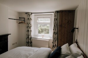 A bedroom with a bed, a radiator, a window with curtains, a wooden wall, and a black lamp. - Charming 2-bedroom cottage with countryside views (Weymouth)