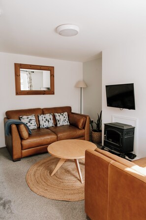 Ein Wohnzimmer mit braunem Ledersofa, einem runden Couchtisch, einem an der Wand befestigten Fernseher und einem Kaminofen.